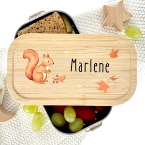 Pu&ograve; includere: Un portapranzo in legno con un'illustrazione di scoiattolo e il nome "Marlene" in nero. Il coperchio presenta foglie arancioni e venature chiare del legno. Il portapranzo contiene un panino e uva.