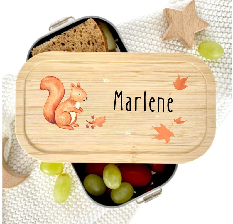 Pu&ograve; includere: Un portapranzo in legno con un'illustrazione di scoiattolo e il nome "Marlene" in nero. Il coperchio presenta foglie arancioni e venature chiare del legno. Il portapranzo contiene un panino e uva.