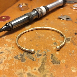 May include: A silver-colored open cuff bracelet with rounded ends, resting on a textured, orange-brown surface. A silver tool and a gold ring are in the background, suggesting jewelry making. The bracelet's design is simple and elegant.