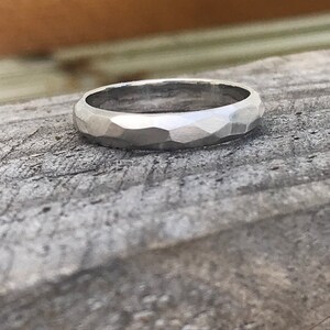 May include: A silver ring with a faceted, hammered texture. The ring is set against a weathered, gray wooden surface, with the ring's surface catching the light.