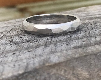 May include: A silver ring with a faceted, hammered texture. The ring is set against a weathered, gray wooden surface, with the ring's surface catching the light.