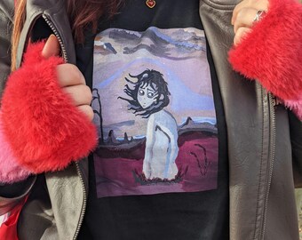 Może przedstawiać: Czarny T-shirt z kolorowym nadrukiem graficznym przedstawiającym kobietę z długimi ciemnymi włosami stojącą w krajobrazie. Kobieta ma na sobie białą sukienkę i ma smutny wyraz twarzy. Krajobraz jest połączeniem kolorów fioletowego, różowego i niebieskiego. T-shirt jest noszony przez osobę w czerwonej kurtce z futra i złotym naszyjnikiem z czerwonym sercem.