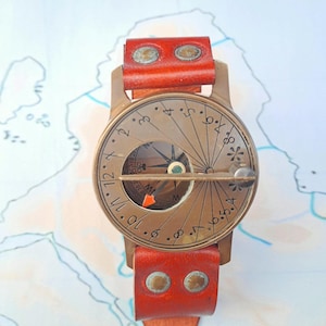 May include: A brass sundial watch featuring a circular face with engraved numbers from 1 to 12, a central gnomon, and an integrated compass. The timepiece is attached to a reddish-brown leather strap secured with two metallic rivets at each lug.