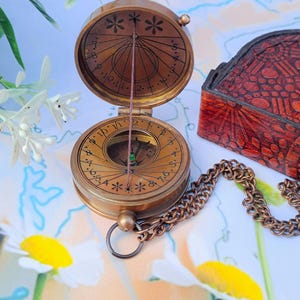 Puede incluir: Una brújula de reloj de sol de latón con tapa abatible, que presenta marcas de hora grabadas y un gnomon de hilo vertical fino. El dispositivo circular incluye una brújula magnética en el centro y una cadena de latón a juego.