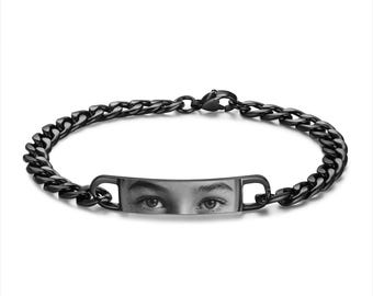 May include: Black chain bracelet with a rectangular tag featuring a black and white photo of a person's eyes.