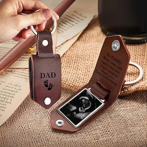 May include: A brown leather keychain with a silver key ring. The keychain has the word "DAD" and baby footprints printed on it. The keychain opens to reveal a black and white sonogram image. The text "To Daddy" is also printed on the keychain.
