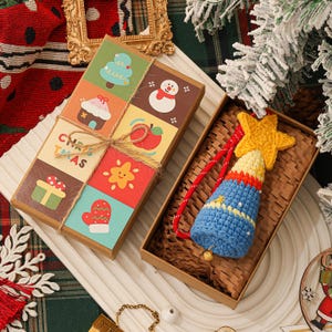 May include: A rectangular cardboard gift box with a grid of colorful Christmas-themed illustrations and twine, containing a crocheted bell ornament with a yellow star topper, a blue body, and a small gold-toned metal bell at the base.