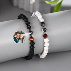 May include: Two beaded bracelets with a photo charm. One bracelet is black with brown beads and the other is white with brown beads. Both bracelets have a silver charm with a black stone.