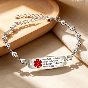Peut inclure: Un bracelet d'alerte médicale argenté composé d'une chaîne à maillons en forme de cœur et d'une plaque d'identification centrale incurvée. La plaque présente un symbole rouge de l'Étoile de Vie et le texte gravé : Anna, Type 2 Diabetes, Allergic to Penicillin, ICE: George: 123-456-789, Kate: 098-765-4321.