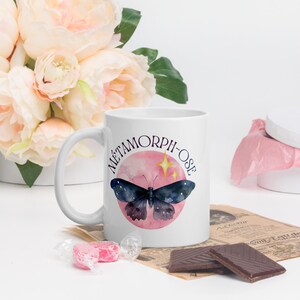 May include: White ceramic mug with a pink and blue butterfly design and the text "METAMORPH-OSE" in purple lettering.