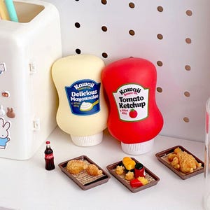 May include: A still-life image featuring miniature food items and condiments. Two squeeze bottles of "Kawaii Sauce Co." mayonnaise and ketchup are next to small trays of food. A tiny Coca-Cola bottle is also present, suggesting a playful, food-themed collection.