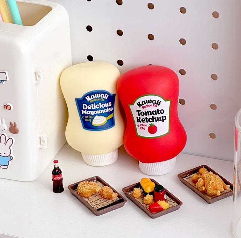May include: A still-life image featuring miniature food items and condiments. Two squeeze bottles of "Kawaii Sauce Co." mayonnaise and ketchup are next to small trays of food. A tiny Coca-Cola bottle is also present, suggesting a playful, food-themed collection.