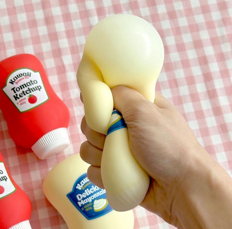 May include: A hand squeezing a large, soft, yellow squishy toy shaped like a mayonnaise bottle. The toy is held near a red ketchup bottle and a yellow mayonnaise bottle, both with text labels. The background is a pink and white checkered pattern.