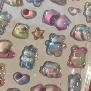 May include: A sheet of iridescent stickers featuring various shapes, including bears, stars, hearts, and geometric forms. The stickers have a translucent, jelly-like appearance with colors ranging from pink and blue to yellow and purple.