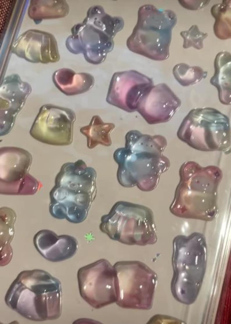 May include: A sheet of iridescent stickers featuring various shapes, including bears, stars, hearts, and geometric forms. The stickers have a translucent, jelly-like appearance with colors ranging from pink and blue to yellow and purple.