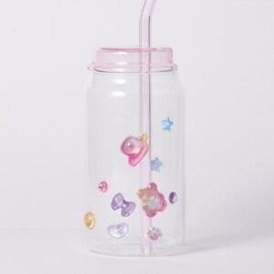 May include: A clear glass tumbler with a pink lid and straw, adorned with colorful, iridescent, and cartoon-like embellishments. The decorations include stars, hearts, bows, and a bear. The tumbler is designed to resemble a soda can.