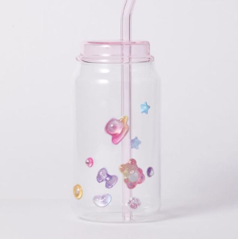 May include: A clear glass tumbler with a pink lid and straw, adorned with colorful, iridescent, and cartoon-like embellishments. The decorations include stars, hearts, bows, and a bear. The tumbler is designed to resemble a soda can.