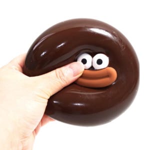 May include: A brown, rounded stress ball with a cartoon face featuring large white eyes and a brown mouth. The stress ball is being squeezed by a hand, showing the malleable nature of the toy.