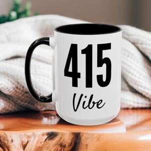 May include: White ceramic mug with a black handle and black rim. The mug has black text that says "415 Vibe" on it.