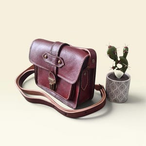 Peut inclure: Sac bandoulière en cuir bordeaux avec fermeture à rabat et bandoulière réglable. Le sac présente des coutures blanches et un fermoir en métal. Un petit cactus en pot avec un pot géométrique est à côté du sac.