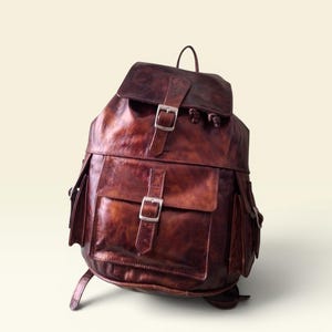 Peut inclure: Un sac à dos en cuir marron foncé doté d'un rabat supérieur avec une boucle argentée, d'une grande poche avant avec une boucle assortie et de deux poches latérales. Le cuir présente une finition patinée avec des variations de tons.