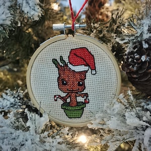 Könnte beinhalten: Ein Weihnachtsbaumschmuck mit einem Kreuzstich-Design eines grünen, braunen und roten Charakters mit einem roten Hut. Der Charakter hält in jeder Hand einen Zuckerstock und steht in einem grünen Topf. Der Schmuck hängt an einer roten und weißen Schnur.