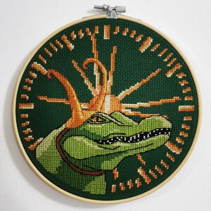 Op de afbeelding: Een geborduurd ontwerp van een krokodil met een doornenkroon, geborduurd in groen en oranje op een groene achtergrond. Het ontwerp is ingelijst in een bruine houten ring.