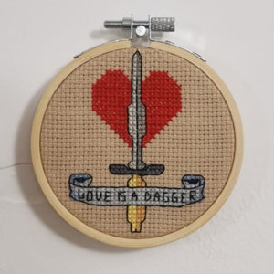 May include: A cross-stitch embroidery hoop with a red heart pierced by a silver dagger. The hoop is made of natural wood and the embroidery features the text "Love is a dagger".
