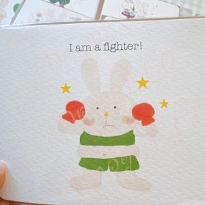 Puede incluir: Tarjeta de felicitación con un conejo boxeador de dibujos animados con guantes rojos y pantalones cortos verdes, rodeado de estrellas amarillas. El texto "I am a fighter!" está impreso encima del conejo. Otras tarjetas con ilustraciones de conejos son visibles en el fondo.