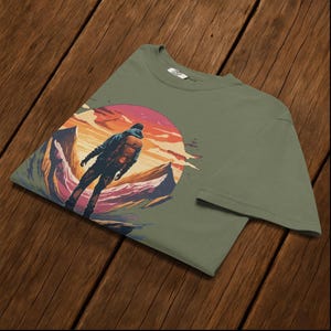 May include: Olive green t-shirt with a graphic of a person with a backpack, standing before a mountain range and a sunset. The design uses shades of orange, pink, and purple. The shirt is made of a soft material.