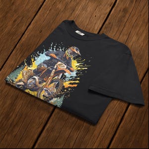 May include: Black t-shirt featuring a motocross rider on an orange dirt bike. The graphic includes colorful splashes of yellow, blue, and green, suggesting speed and action. The rider is wearing a helmet and protective gear.