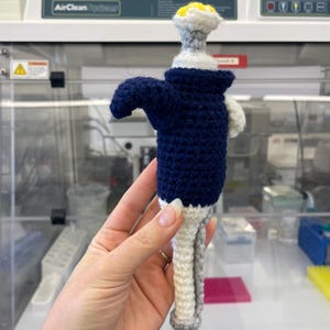 May include: Crocheted pipette in navy blue, white, gray, and yellow. The handmade lab tool features a navy blue body, white and gray handle, and a yellow top. The pipette is held in front of a laboratory workstation.