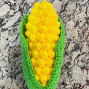 Puede incluir: Una mazorca de maíz tejida a crochet con cáscaras verdes. El maíz es amarillo y las cáscaras son verdes.
