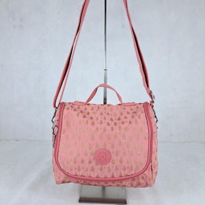 May include: A pink shoulder bag with a gold teardrop pattern. The bag has a top handle and a long, adjustable strap. A small, round logo is centered on the front. The bag is displayed on a metal stand.
