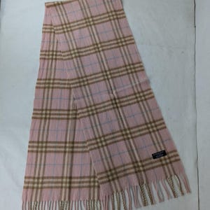 May include: A pink scarf with a classic plaid pattern in brown and light blue. The scarf has a fringed edge and a small label with text. The scarf is made of a soft material.
