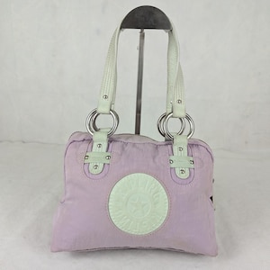 Peut inclure: Un sac à main lavande avec un patch circulaire vert clair avec une étoile et du texte. Le sac a deux poignées blanches avec des anneaux argentés et des accents vert clair. Le sac est présenté sur un support métallique.