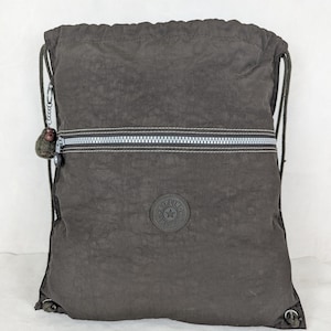 May include: A dark gray drawstring backpack with a silver zipper and a small round logo. The backpack has a textured appearance and a small monkey keychain attached to the drawstring. The bag is made of a lightweight material.