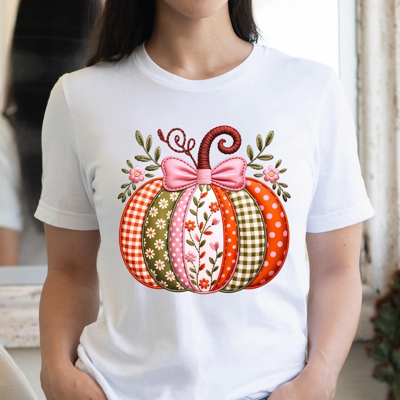 May include: White t-shirt with a decorative pumpkin design. The pumpkin is made up of different patterns, such as gingham, polka dots, and floral prints, in orange, pink, green, and red. A pink bow and leafy details complete the design.