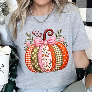 May include: A grey t-shirt with a pumpkin design. The pumpkin is segmented with patterns like orange polka dots, green gingham, and floral embroidery. A pink bow and leaves adorn the pumpkin. The design is autumnal in theme.