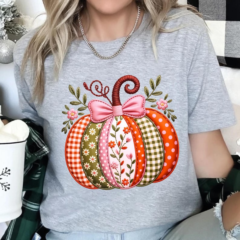 May include: A grey t-shirt with a pumpkin design. The pumpkin is segmented with patterns like orange polka dots, green gingham, and floral embroidery. A pink bow and leaves adorn the pumpkin. The design is autumnal in theme.