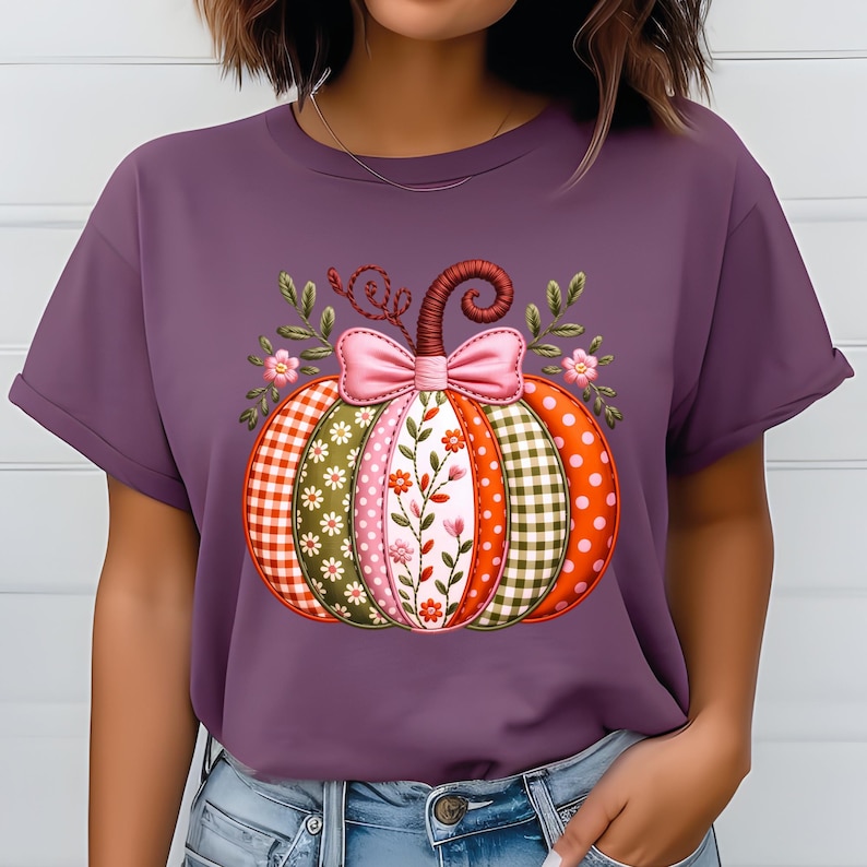 May include: A purple t-shirt with a decorative pumpkin design. The pumpkin is made up of segments with different patterns, such as gingham, polka dots, and floral prints. A pink bow and floral details decorate the pumpkin, creating a seasonal autumn theme.