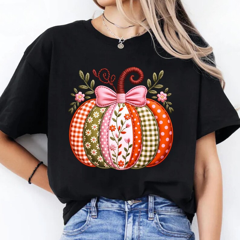 May include: Black t-shirt with a decorative pumpkin design. The pumpkin is segmented with patterns like gingham, polka dots, and floral motifs. A pink bow and green leaves are at the top of the pumpkin.