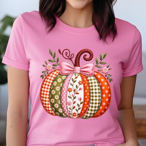 May include: A pink t-shirt with a decorative pumpkin design. The pumpkin is divided into sections with patterns like gingham, polka dots, and floral prints. A pink bow and leafy details are at the top, creating a seasonal autumn theme.