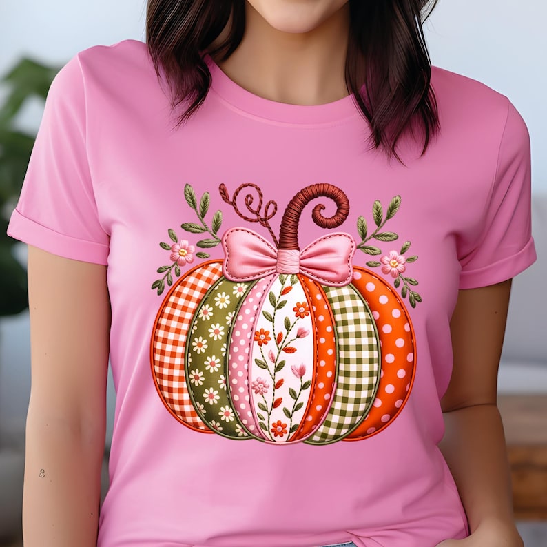 May include: A pink t-shirt with a decorative pumpkin design. The pumpkin is divided into sections with patterns like gingham, polka dots, and floral prints. A pink bow and leafy details are at the top, creating a seasonal autumn theme.