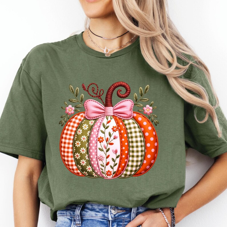 May include: An olive green t-shirt with a pumpkin design. The pumpkin is made up of segments with different patterns, including orange polka dots, red and white gingham, and floral prints. A pink bow and floral accents complete the autumn-themed graphic.