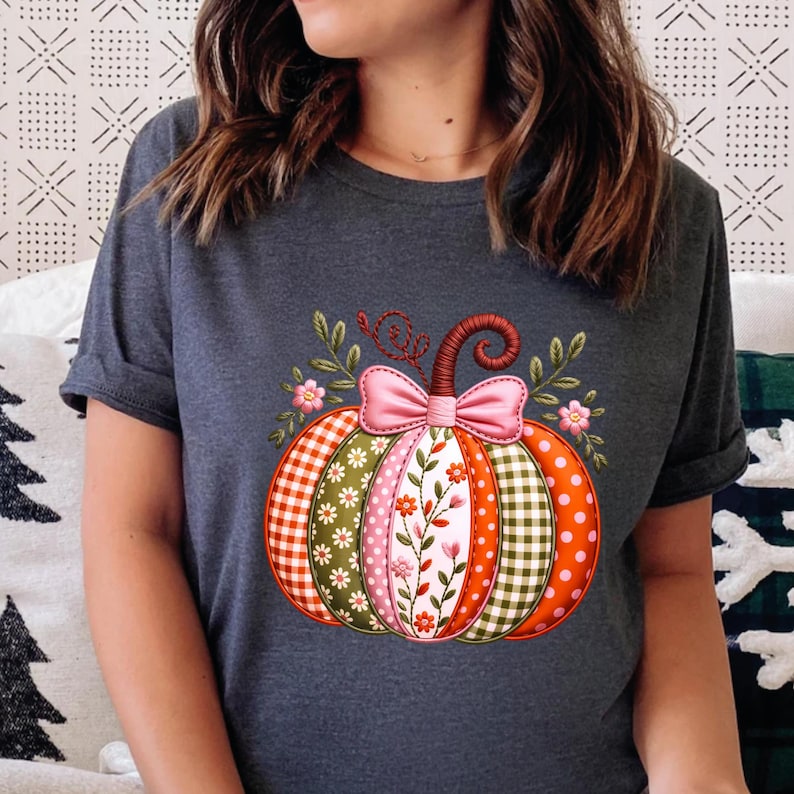 May include: Dark grey t-shirt with a vibrant pumpkin design. The pumpkin is decorated with a mix of patterns, such as gingham, polka dots, and floral motifs. A pink bow and floral details embellish the top, creating a festive autumn theme.