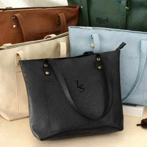 May include: An assortment of leather tote bags in various colors: brown, cream, green, light blue, and black. Each bag features a zipper closure, handles, and some have personalized initials. Sunglasses and a candle are also present.