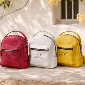 May include: Three small backpacks in magenta, white, and yellow with a pebbled texture. Each bag features a top handle, a front zippered pocket, and black metal hardware. The magenta bag has the letters KN, the white bag says DOMINIKA, and the yellow bag has AK.