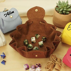 May include: A selection of leather dice bags and a brown leather dice tray. The bags are in various colours, including white, blue, yellow, pink, and purple. Each bag has a name printed on it. Dice are scattered around the tray and bags.