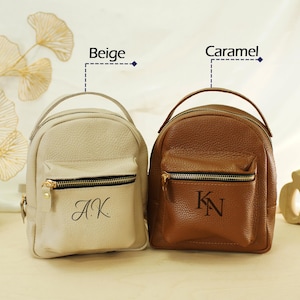 May include: Two small pebbled leather backpacks with top handles and front zippered pockets. The beige backpack features the monogram A.K., while the caramel backpack displays the monogram K.N. Both bags have gold-toned hardware and zippers.